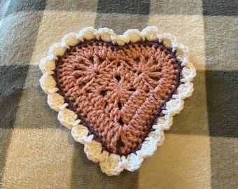 May include: A heart-shaped crochet coaster featuring a dusty rose center, a dark brown border, and a ruffled white edge. The piece is constructed with visible yarn stitches in a decorative pattern, creating a textured, layered appearance.