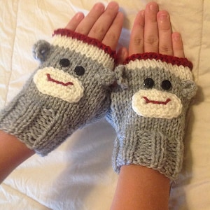 May include: A pair of gray knitted fingerless gloves with monkey faces. The gloves have white faces, black button eyes, and red smiles. The cuffs are white with a red trim. The gloves are designed to cover the hand up to the knuckles.