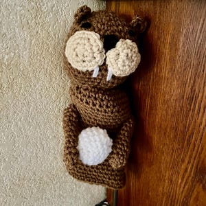 May include: A brown crocheted beaver figure featuring two large beige circular cheeks, a small black nose, two white protruding teeth, and a white crocheted ball held in its front paws. The figure has small rounded ears and a textured yarn finish.