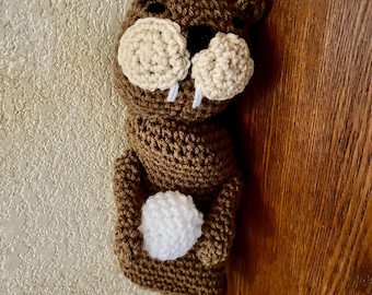 May include: A brown crocheted beaver figure featuring two large beige circular cheeks, a small black nose, two white protruding teeth, and a white crocheted ball held in its front paws. The figure has small rounded ears and a textured yarn finish.