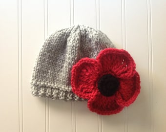 May include: A light gray knitted beanie hat featuring a large, crocheted red poppy flower with a dark black center attached to the side. The hat is constructed with a ribbed brim and a smooth stockinette stitch pattern on the main body of the cap.