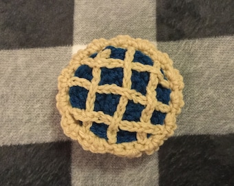 May include: A round, crocheted pie with a deep blue filling and a tan, lattice-style crust. The top of the pie features a woven, crisscross pattern made of yarn, with a scalloped, textured edge that mimics the appearance of a traditional baked pastry.