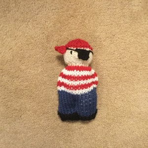May include: A knitted toy pirate with a red and white striped shirt, blue pants, and a red bandana. The toy has a black eye patch.