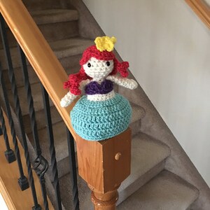 Puede incluir: Una mu&ntilde;eca de sirena de crochet con cabello rojo, un top morado y una cola turquesa. La mu&ntilde;eca lleva una corona amarilla y est&aacute; sentada en una barandilla de madera.