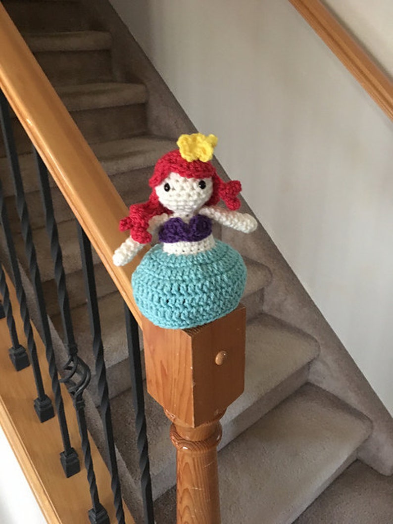 Puede incluir: Una mu&ntilde;eca de sirena de crochet con cabello rojo, un top morado y una cola turquesa. La mu&ntilde;eca lleva una corona amarilla y est&aacute; sentada en una barandilla de madera.