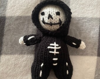 May include: A black knitted skeleton doll with a white skull and ribcage design. The doll is wearing a black hooded robe.