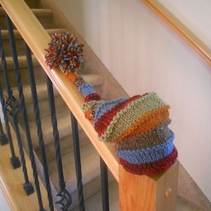May include: A colorful knitted hat with a pom-pom on top, hanging on a wooden handrail of a staircase.