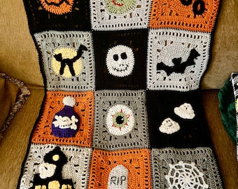 May include: A handmade crochet blanket featuring a grid of twelve square panels with Halloween motifs including a pumpkin, a zombie face, the word BOO, a black cat, a skull, a bat, a potion bottle, an eyeball, ghosts, a house, a tombstone with RIP, and a spiderweb.