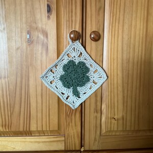 Peut inclure: Un dessous de plat carré au crochet avec un motif de trèfle vert au centre. Le dessous de plat est beige clair avec un bord festonné et est suspendu à une porte d'armoire en bois. Le trèfle est d'une nuance de vert plus foncée.
