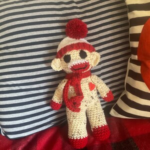 May include: A crocheted toy monkey wearing a red and white hat with a red pom-pom on top. The monkey is wearing a red scarf and has a red heart on its chest.