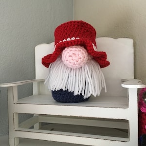 May include: A crocheted gnome with a red and white hat, a pink nose, and a white beard. The gnome is sitting on a white bench.