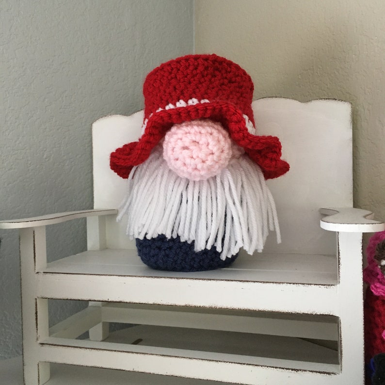 May include: A crocheted gnome with a red and white hat, a pink nose, and a white beard. The gnome is sitting on a white bench.