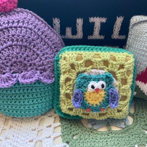 May include: A crocheted owl toy with yellow, green, and purple yarn. The owl is sitting inside a crocheted square with green and yellow yarn. The square is sitting on a white crocheted doily.