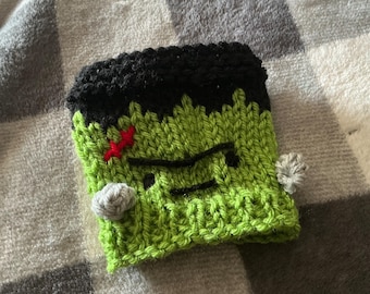 May include: A lime green knitted cup cozy featuring a black hair section at the top, black embroidered eyes and a mouth, a small red stitched scar on the forehead, and two small grey circular accents on the sides resembling bolts on a monster.