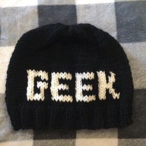Può includere: Berretto nero a maglia con la parola "GEEK" in lettere bianche a maglia.