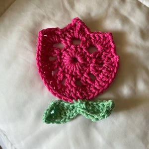 May include: A handmade crochet flower in bright pink with a green leaf. The flower has a detailed openwork design. The crochet flower is likely used for embellishments or decorative purposes. The item is set against a white background.