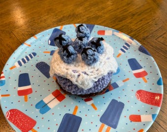 May include: A crocheted blueberry muffin toy featuring a light purple base, a white crocheted topping, and several small blue crocheted blueberries with black embroidered details on top, resting on a light blue plate with popsicle patterns.