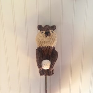 May include: Knitted golf club head cover shaped like a bear holding a golf ball. The bear is brown and tan with black eyes.