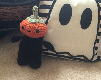 May include: A crocheted pumpkin-shaped plush toy with a black body and orange pumpkin head. The toy is standing next to a white pillow with a black ghost design.