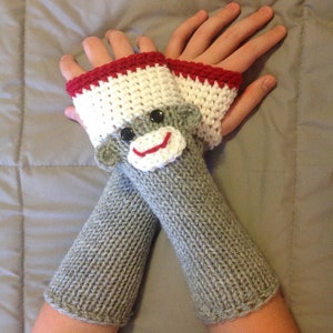 May include: Hand-knitted gray fingerless gloves with a monkey face design. The gloves feature white and red crochet accents at the wrist and a monkey face with black button eyes and a red smile. A fun and cozy accessory.