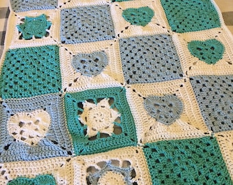 May include: A handmade crochet blanket featuring a patchwork design. The blanket incorporates squares in white, teal, and light blue, with heart and floral motifs. The blanket is displayed on a checkered surface.