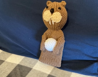 May include: A brown knitted beaver toy featuring a round head with small ears, black embroidered eyes, a black nose, and two white teeth. The beaver has a light cream-colored muzzle and is holding a small, round, white crocheted ball in its arms.