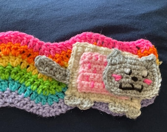 May include: A crocheted headband featuring a vibrant rainbow-colored wavy pattern. Attached to the front is a gray cat figure with pink cheeks, resting on a cream-colored rectangular base with a pink center, all crafted from yarn.