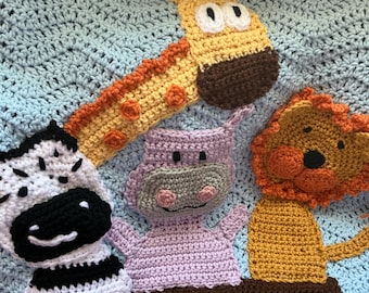 May include: A light blue crocheted blanket featuring appliqued yarn animals including a yellow giraffe, a black and white zebra, a lavender hippo, and an orange lion, all positioned above a dark green crocheted border representing grass.