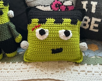 May include: A square, lime green crochet pillow featuring a black hair section, white eyes with black pupils, a small black mouth, and a red stitched scar on the forehead. The pillow has small grey side protrusions and a textured yarn construction.