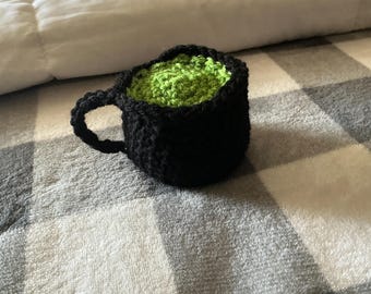 May include: A small, handmade crochet mug featuring a solid black exterior and a textured, bright lime green interior. The item has a rounded shape with a single black handle attached to the side, crafted from yarn with visible stitching throughout.