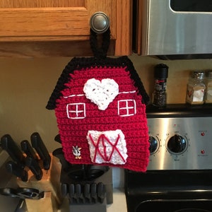 May include: A red crocheted pot holder shaped like a barn with a white heart, white windows, and a white 'XO' on the door. A small white crocheted chicken is on the side of the barn.