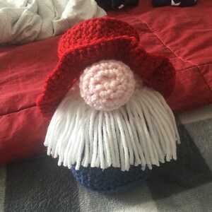May include: A crocheted gnome with a red hat, white beard, and blue body. The gnome is sitting on a patterned surface.