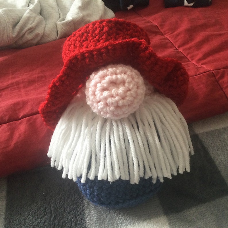 May include: A crocheted gnome with a red hat, white beard, and blue body. The gnome is sitting on a patterned surface.