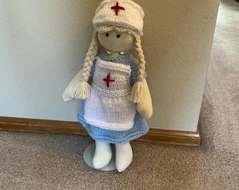 May include: A fabric doll dressed as a nurse, featuring a light blue knitted dress, a white knitted apron with a red cross, and a matching white nurse cap with a red cross. The doll has yarn hair styled in two long braids and a plain face.