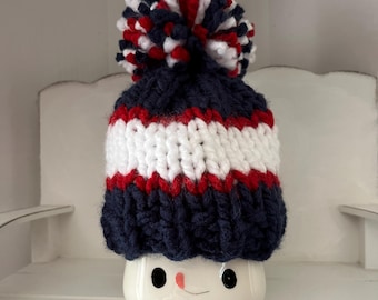 May include: A white ceramic mug shaped like a snowman face, featuring two black eyes and a small orange carrot nose. The mug is topped with a chunky knit beanie in navy blue, white, and red, finished with a large matching multi-colored pom-pom.