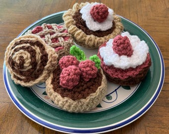 May include: A collection of five crocheted play food items arranged on a plate. The set includes a cinnamon roll, a lattice-top pie, and three small tarts topped with white yarn frosting and red yarn berries or green yarn leaves.