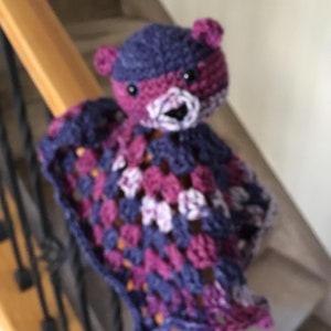 May include: A purple and blue crocheted blanket with a small crocheted bear attached. The bear has a pink nose and black eyes.