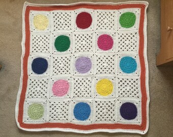 May include: A white crocheted blanket with a red border and colorful circles in the center. The circles are in shades of red, green, blue, yellow, pink, and purple.