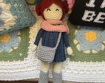 May include: A handmade crochet doll with red hair styled in two buns, wearing a dark blue dress, a light pink scarf, and gray boots. The doll has a cream-colored face with embroidered closed eyes and a smile, and carries a small gray crochet bag.