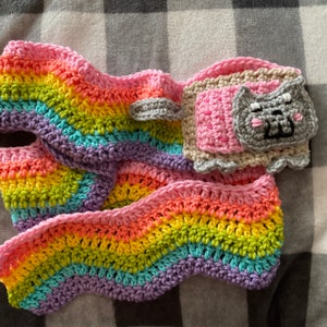 Puede incluir: Bufanda de ganchillo hecha a mano en colores del arcoíris, con tonos rosa, amarillo, verde, azul y morado. La bufanda tiene una aplicación de cara de gato y un pequeño bolsillo. Con borde ondulado.