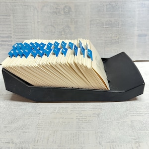 May include: A black plastic rotary card file holder containing a series of off-white index cards. The cards are organized with blue alphabetical tab dividers, featuring letters from A to W, arranged in a slanted, fanned-out position within the base.