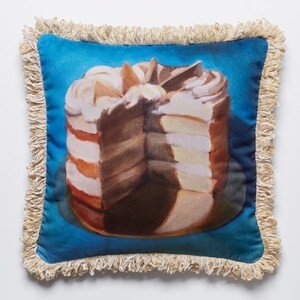 Könnte beinhalten: Ein quadratisches Kissen mit blauem Hintergrund und einer detaillierten Illustration eines mehrschichtigen Kuchens mit weißem Frosting und Schokoladenfüllung. Das Kissen hat einen Fransenrand.