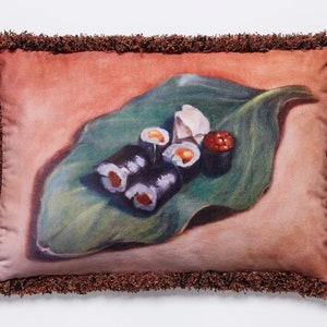 May include: Rectangular decorative pillow with a sushi design. The pillow features a brown fringe border and a print of sushi rolls, ginger, and roe on a large green leaf against a peach background.