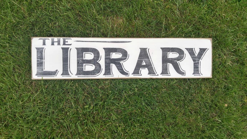 Op de afbeelding: Een wit houten bord met zwarte letters waarop "THE LIBRARY" staat. Het bord ligt op een bed van groen gras.