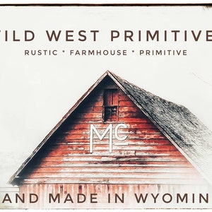 Op de afbeelding: Een rode houten schuur met een wit "MC"-logo aan de zijkant. De schuur staat tegen een witte achtergrond met de tekst "WILD WEST PRIMITIVES RUSTIC * FARMHOUSE * PRIMITIVE HAND MADE IN WYOMING" boven de schuur.