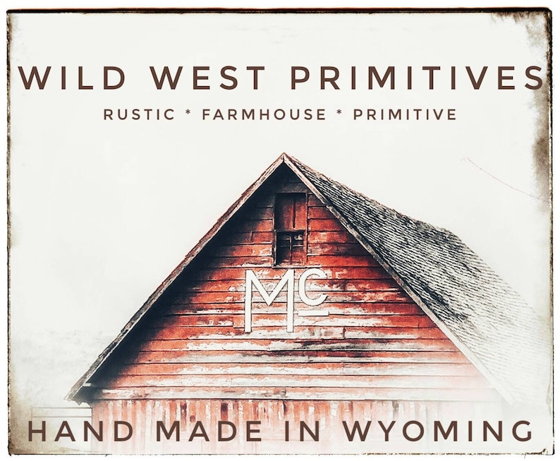 Op de afbeelding: Een rode houten schuur met een wit "MC"-logo aan de zijkant. De schuur staat tegen een witte achtergrond met de tekst "WILD WEST PRIMITIVES RUSTIC * FARMHOUSE * PRIMITIVE HAND MADE IN WYOMING" boven de schuur.