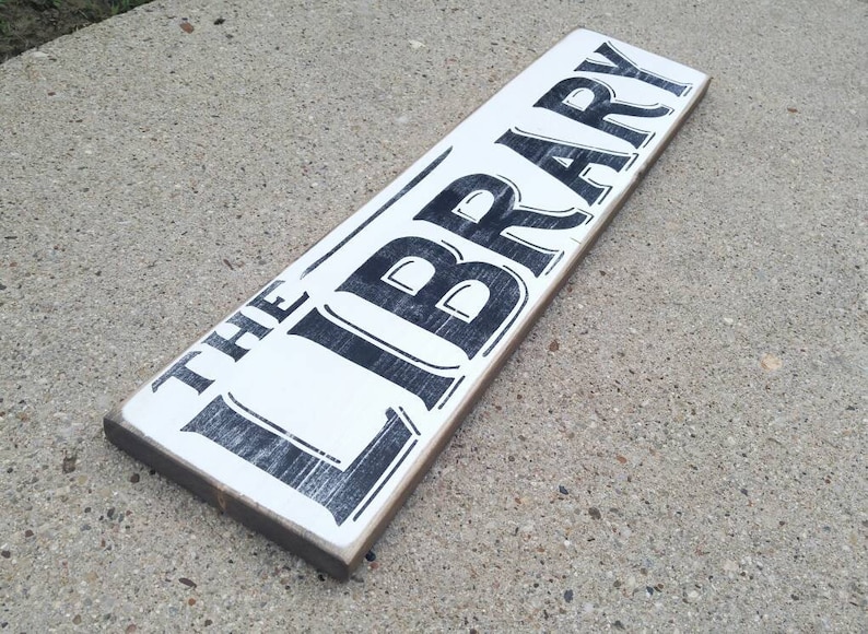Op de afbeelding: Een wit houten bord met zwarte letters waarop "THE LIBRARY" staat. Het bord heeft een verweerde uitstraling en is perfect om een ​​rustieke charme aan uw interieur toe te voegen.
