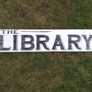 Op de afbeelding: Een wit houten bord met zwarte letters waarop "THE LIBRARY" staat. Het bord ligt op een bed van groen gras.