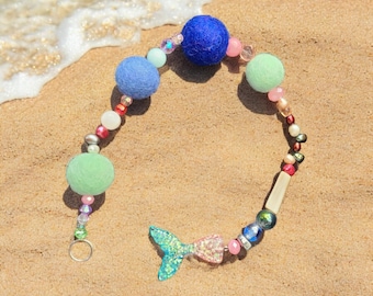 Könnte beinhalten: Eine bunte Perlenkette mit Filzkugeln in Blautönen, Grün- und Rosatönen. Die Kette hat einen Meerjungfrauen-Schwanz-Anhänger und einen silbernen Ring.