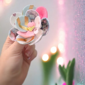 May include: A handmade felt flower with gold accents and a pink center. The flower is made of multiple layers of felt in different colors and patterns.